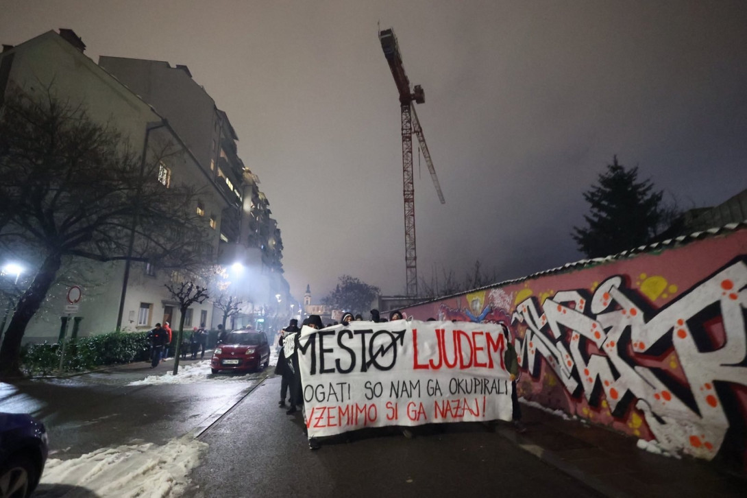Večerna fotografija Trubarjeve ulice, viden je grafitiran zid tovarne rog na desni, skupina temno oblečenih oseb drži transparent z napisom MESTO LJUDEM / Bogati so nam ga okupirali / Vzemimo ga nazaj. V ozadju se vzpenja žerjav